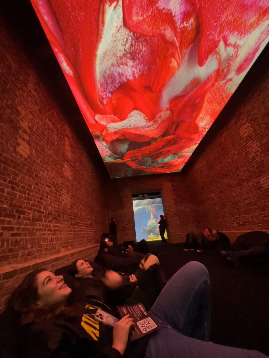 People laying on the ground staring up at ceiling screen with red and yellow video