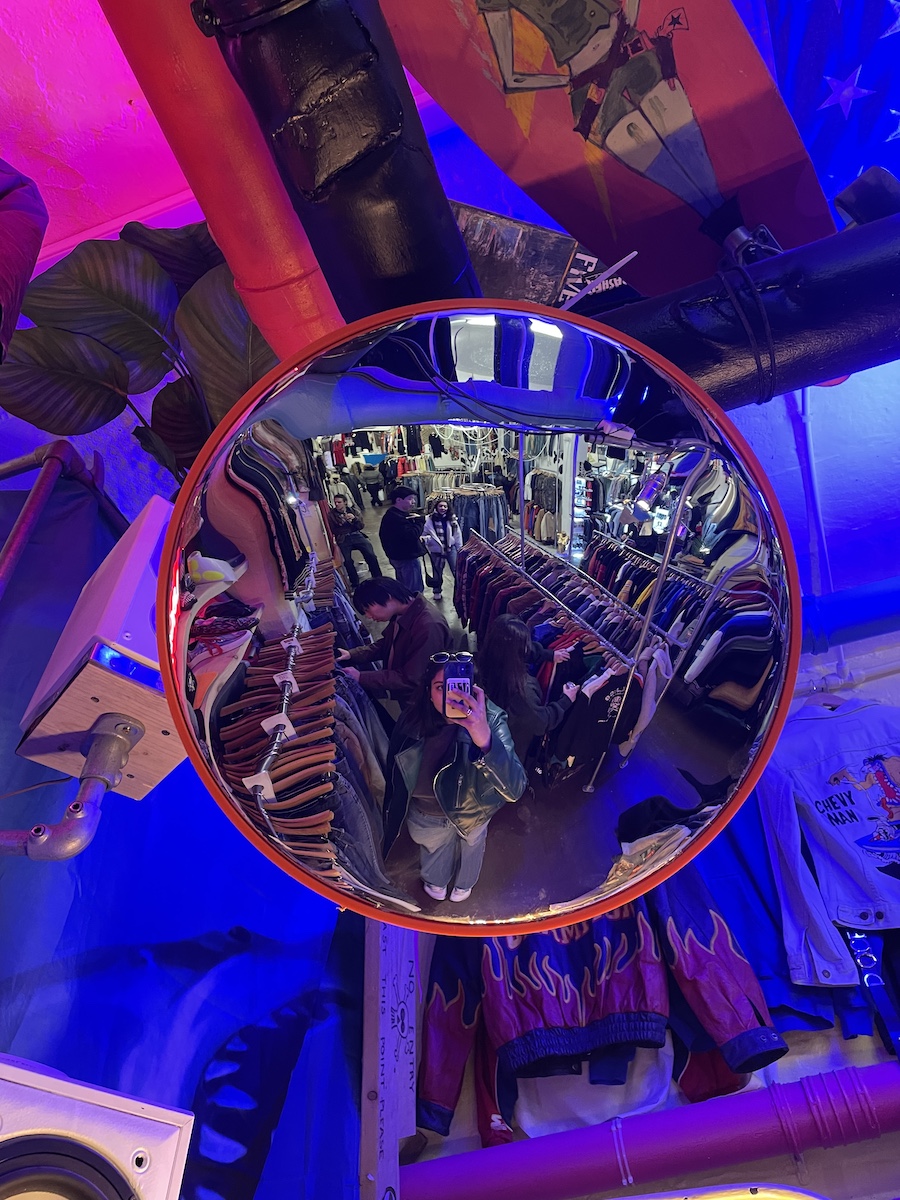 Girl taking photo through wide mirror, with rows of clothing racks in background in blue lighting