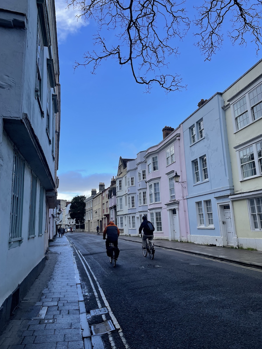 Two men's backs biking on street with pale, colorful houses